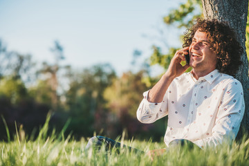 Attractive happy male with curly hair wearing trendy shirt calling his girlfriend to tell that his wainting her in the park. Copy space for technology and communication advertising. People, lifestyle
