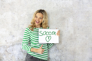 young pretty woman with striped pullover holding checkered paper block in the camera with the word soccer written on it