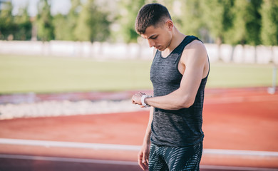 Caucasian athlete guy standing outdoors in stadium using a smartwatch to monitor his progress. Male athlete resting and checking his performance on fitness smartwatch device. People and sport concept