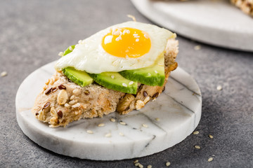 Healthy breakfast with open sandwich with fresh avocado and fried quail egg on small marble board on gray background.