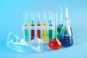Chemical glassware with colorful samples and lab goggles on color background
