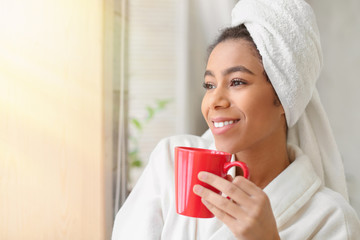 Beautiful African-American woman in bathrobe drinking tea at home