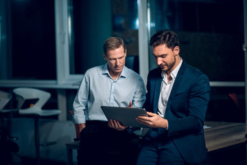 Two office collegues looking at tablet. Handsome businessman wearing suit showing something to his collegue on tablet late in evening. - Powered by Adobe