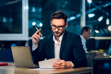 Portrait of businessman working in office. Handsome bearded businessman with glasses sitting at office table looking down and reading paper with pencil in his hand. © Svyatoslav Lypynskyy