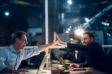 Two businessmen highfiving each other late at night. Two happy male coworkers staying up late in...