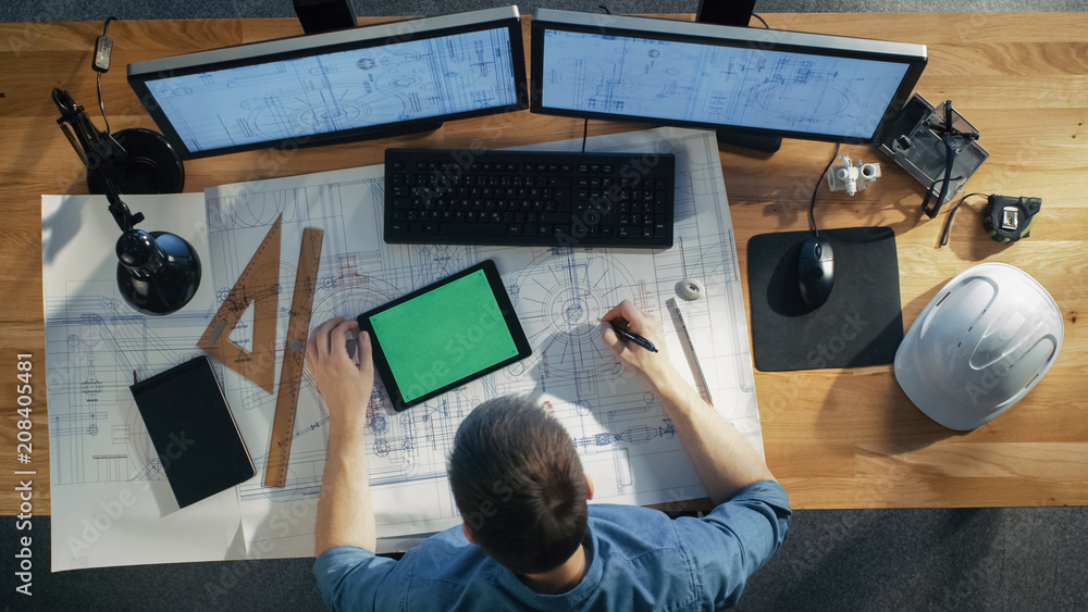 Poster top view of architectural engineer draws on his blueprints, compares with tablet computer with green