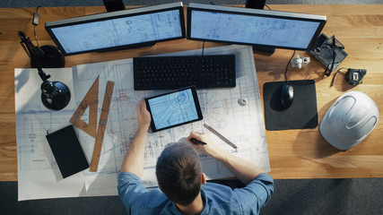 Top View of architectural Engineer Draws on His Blueprints, Compares with Tablet Computer with Green Screen, Using Desktop Computer Also. His Desk is Full of Useful Objects and Evening Sun. - obrazy, fototapety, plakaty