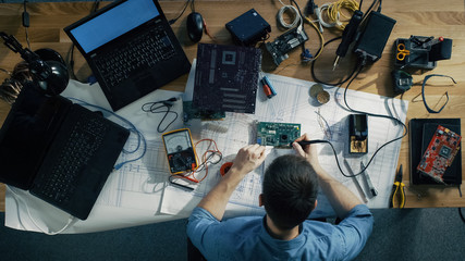 Top View of Young IT Technician Soldering Circuit Board. His Table is Full With Various Computer Parts, Motherboards, Laptops, Cables. Morning Sun Illuminates His Desk.