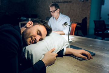 Sleepy businessman resting his head on pillow. Close portrait of smiling handsome man sleeeping on pillow at his office table with his colleague working in background.