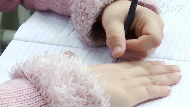 Close-up of children`s hands are writing with a pencil on a white sheet of paper. A little girl is sitting at the table and writing.