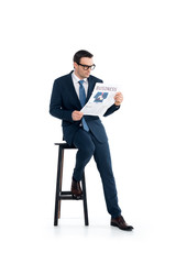 Obraz premium full length view of businessman in suit and eyeglasses sitting on stool and reading newspaper isolated on white