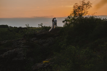 Rear view of romantic wedding couple of groom and bride in gorgeous wedding dress with long loop standing on the rocks in the mountains on sunset. Scenic landscape view