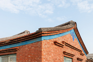 Traditional swallowtail style roof line found in historic villages in China and Taiwan. The building is brick with a decorative blue symbol & band around the building. There is a blue sky and clouds.