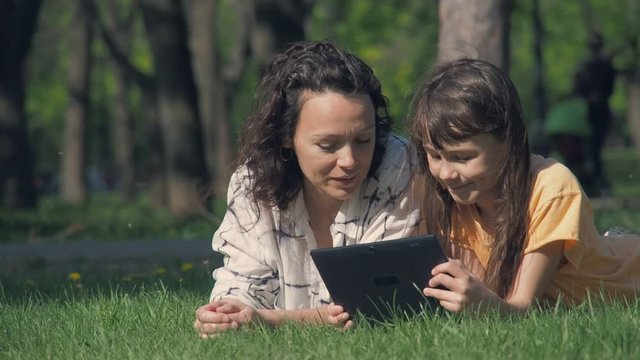 Happy family in the park with a tablet. Mom and daughter on green grass with a gadget.