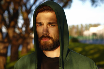attractive pensive young bearded guy wearing green hood pulled over his head, looking at camera...