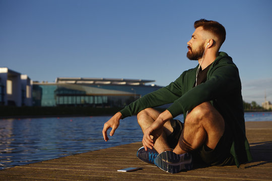 Picture of attractive unshaven young Caucasian guy in running shoes sitting cross legged on wooden paving by lake meditating, listening to calm music using free application on his electronic gadget