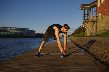 Outdoor summer portrait of confident active young sporty guy with beard exercising outdoors, warming up legs muscles before jogging workout, casting shadow on wooden paving by river or lake