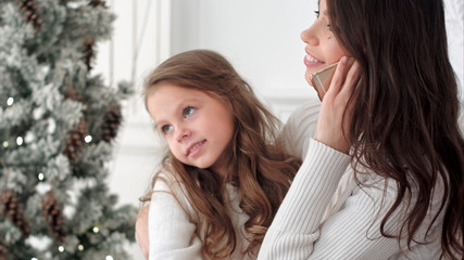 Young mother talking on the phone with slightly bored daughter sitting on her lap next to the xmas tree