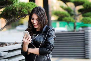 Beautiful young dark-haired woman is texting on her smartphone while sitting the bench in city park