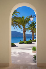 Walkway to the sea. Sunny embankment of Tivat city with green palm trees  is visible through an arch of building. Montenegro, Bay of Kotor (Adriatic Sea)