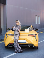 Beautiful blonde young woman with shopping bag near the yellow super car, outdoors
