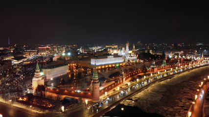 Fototapeta na wymiar Flying above frozen river Moskva and its embankment illuminated with sparkling lanterns. Bright cityscape and Moscow Kremlin at night. Intensive traffic in the streets of Moscow.