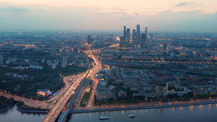 Obraz premium Flying above the wide illuminated city highways and Moskva river. Cityscape at dusk. Moscow-city towers in the evening haze on the horizon.