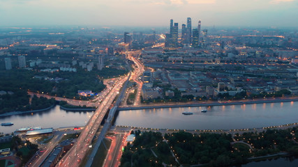 Fototapeta premium Flying above the wide illuminated city highways and Moskva river. Cityscape at dusk. Moscow-city towers in the evening haze on the horizon.
