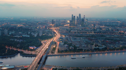 Obraz premium Flying above the wide illuminated city highways and Moskva river. Cityscape at dusk. Moscow-city towers in the evening haze on the horizon.