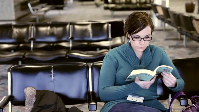 Girl sitting at an airport lobby, waiting for a flight, reading book, smiling at camera.
