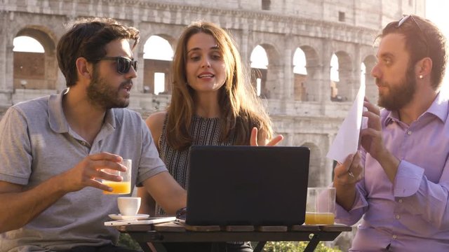 Three young people having job videocall with laptop working outside together pitching a project sitting at bar restaurant table in front of colosseum in rome at sunset