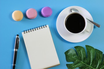 Flat lay photo with notebook , coffee cup Pen, glasses ,Macaroon and Tropical Leave on blue background