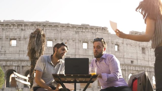 Three young people having job videocall with laptop working outside together pitching a project sitting at bar restaurant table in front of colosseum in rome at sunset