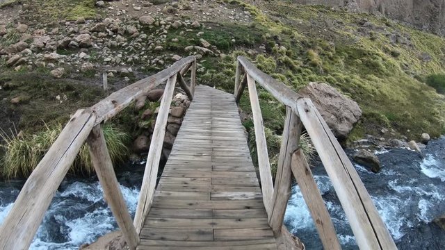 Small wooden pedestrean bridge over Covunco river, in Los Tachos, at Domuyo Volcano. Hot water with geisers on a dark cloudy cold day. Steppe and grasslands. Neuquen, Patagonia Argentina.
