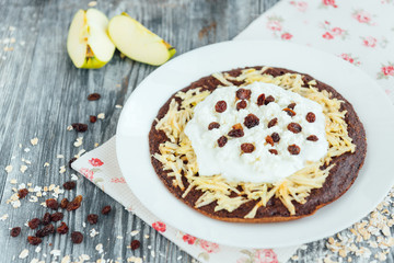 Homemade chocolate oat pancakes with apple, yogurt, cottage cheese, raisin and flowers on grey wooden background. Healthy breakfast or snack.
