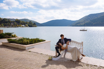 Elegant view of the river embankment. The girl in the wedding dress lay down on a bench on her knees to her husband in a stylish suit and admires the blue sky, the man looks at non-floating yachts.