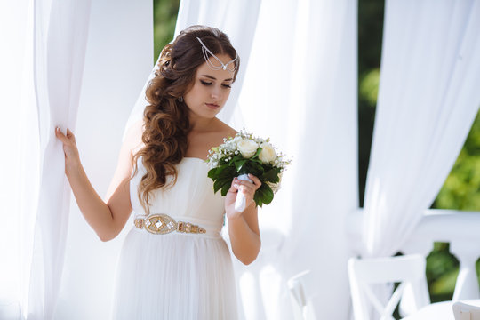 Portrait of the bride in the image of the Greek goddess Aphrodite poses in a cafe decorated for ancient Rome, admires her wedding bouquet, her hair is decorated with beads.