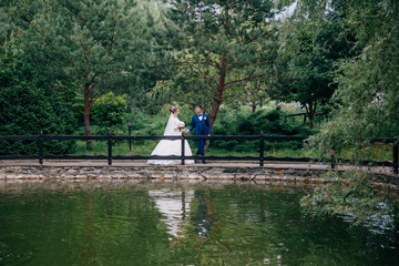 The bride and groom are walking in the green park, the man is leading his wife across the bridge, holding her by the hand. Lovely wedding dress of the bride is reflected in the watery surface.