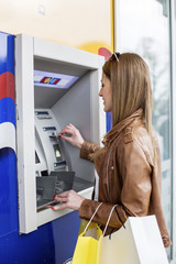 Young beautiful girl in front of ATM with credit card on hand and shopping bags