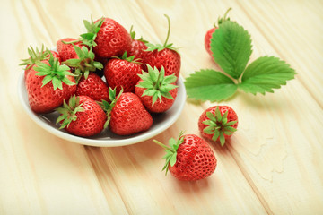Fresh ripe strawberry with green leaves in a white plate on a light wooden background.
