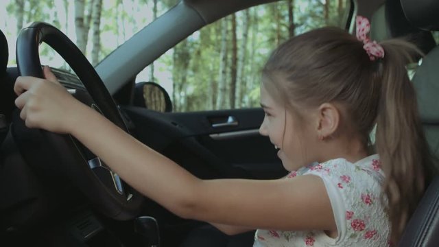 Joyful happy elementary age girl playing in car and pressing car horn on steering wheel while pretending to drive parents car in drivers seat. Smiling child having fun in drivers seat in automobile