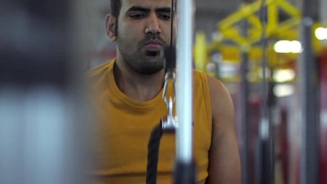 Tilt up of focused Arab man in tank top training on cable triceps pushdown machine with rope attachment, then looking at camera and laughing
