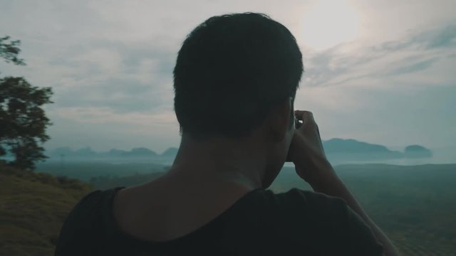 Movement shot of photographer traveler taking photo of beautiful tropical island landscape and cloudy sky while standing on a hilltop during early morning - video in slow motion