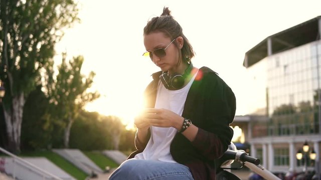 A fashionable, modern girl sits on a moped and texting on her mobile phone. She wears jeans, a white jersey and a black sweater. In sunglasses. Evening dusk. Outdoors