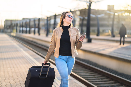 Confident business woman pulling suitcase and holding mobile phone in train station. Smiling lady walking in platform and using smartphone. Female traveler arrived to destination.