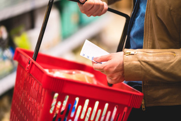 Hand holding shopping list and basket in grocery store aisle. Woman reading paper, shelf in the...