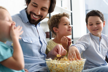 smiling man and little sons eating popcorn while watching film together at home