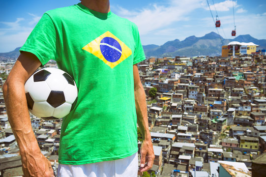 Football player standing in Brazil flag t-shirt holding soccer ball in front of favela slum background in Rio de Janeiro. 