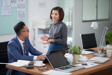 Pretty Asian manager standing at desk of her male colleague and telling him funny story while taking short break from work, interior of open plan office on background