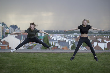 Two young female gymnasts doing acrobatics jumps with cityscape on background